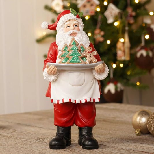 A Santa Claus figurine wearing a red suit and white apron, holding a tray with decorated Christmas trees and gingerbread cookies